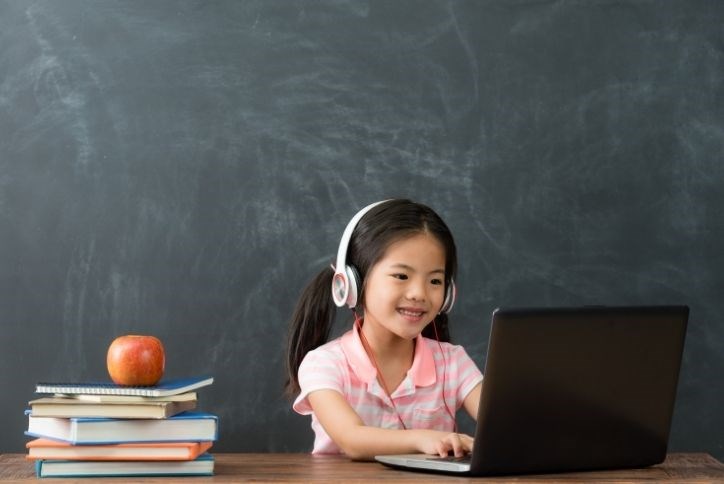 Child working on her laptop for digital learning with headphones on. Books and apple on desk.